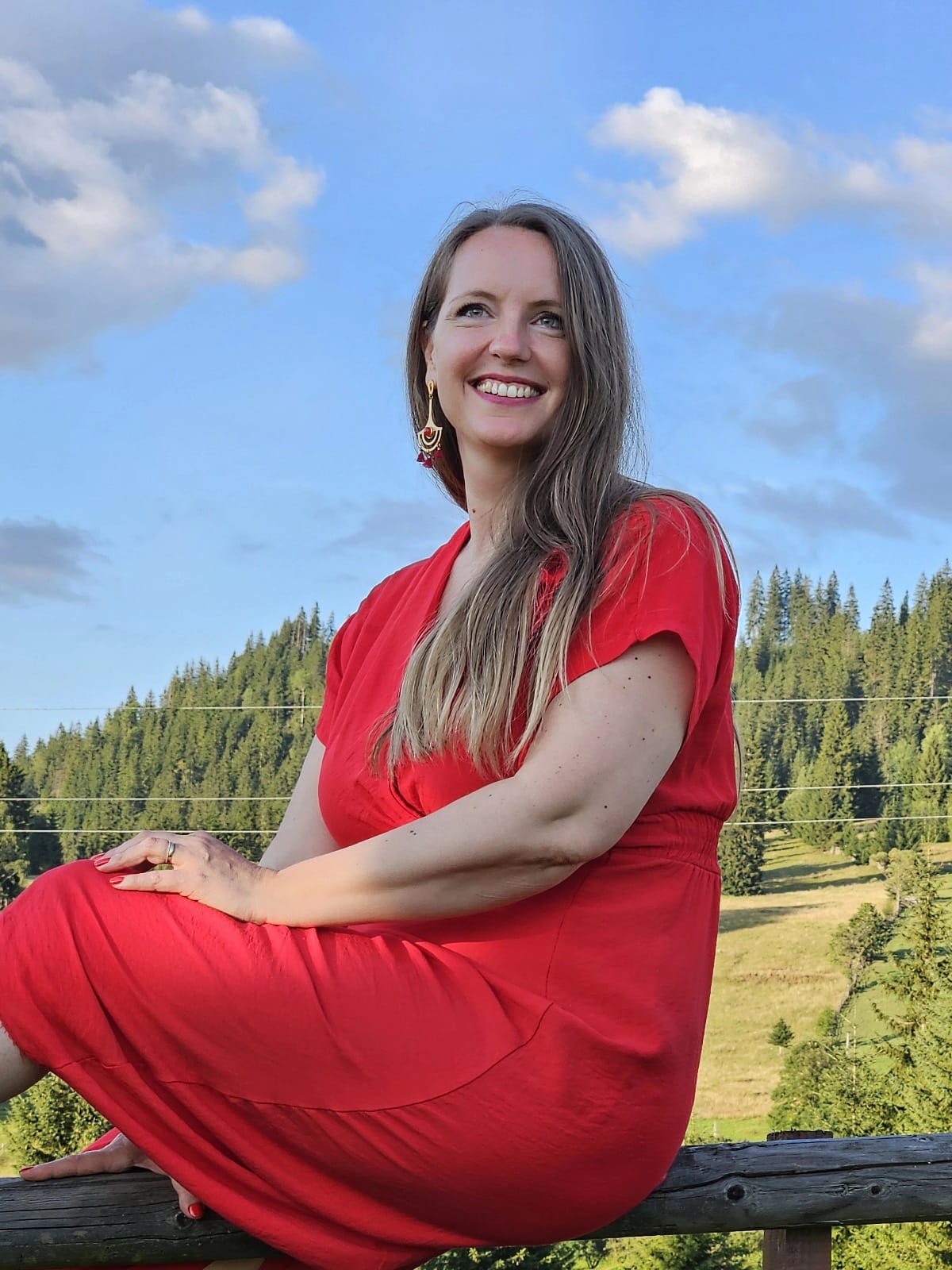 Bianca Varvara, în rochie roșie, așezată pe un gard de lemn cu munții în spate